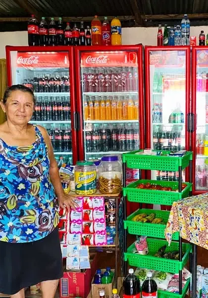 Mujer atendiendo un pequeño negocio con productos en estantes y refrigeradores.